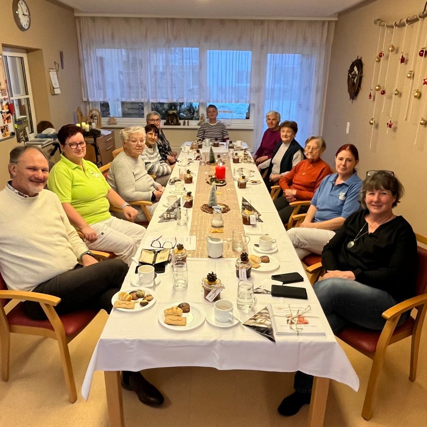 Eine Gruppe älterer Menschen sitzt um einen langen Tisch herum, wahrscheinlich in einem Esszimmer, und isst eine Mahlzeit.