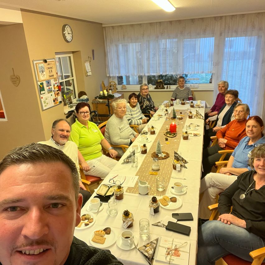 Eine Gruppe älterer Menschen sitzt um einen langen Tisch mit Essen und Getränken, eine Frau in Grün sitzt in der Mitte und ein Mann in Schwarz im Vordergrund.