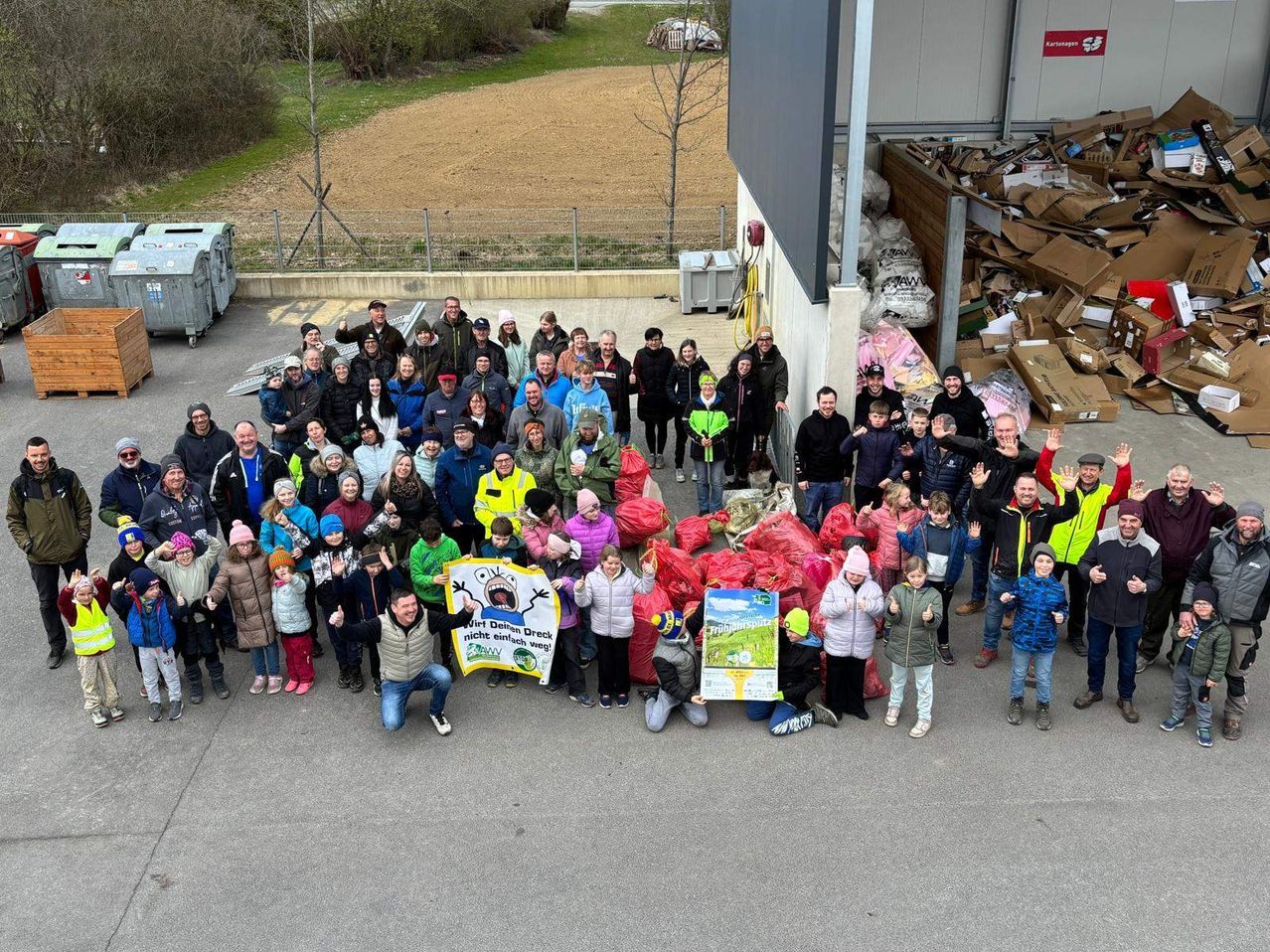 Eine Gruppe von Menschen steht um eine große Ansammlung von Müllsäcken herum, lächelt und winkt für die Kamera. Hinter ihnen befindet sich ein Gebäude mit einem Müllcontainer und Lagerboxen. In der Ferne gibt es ein Grasfeld mit einem Zaun und Bäumen.