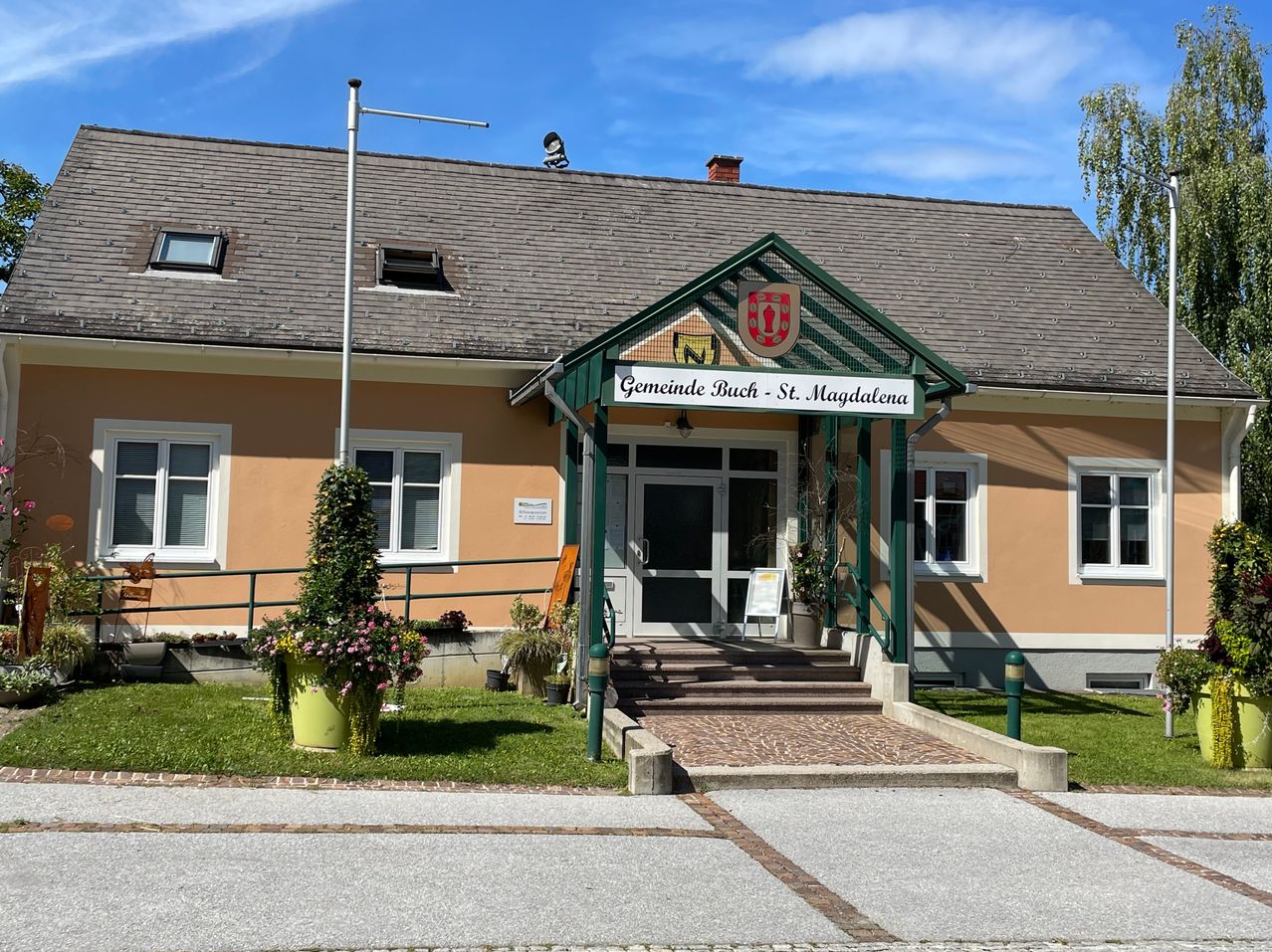 Ein beigefarbenes Gebäude mit einem grünen Vordach über dem Eingang, beschriftet Gemeinde Buch St. Magdalena. Topfpflanzen stehen auf dem Rasen.