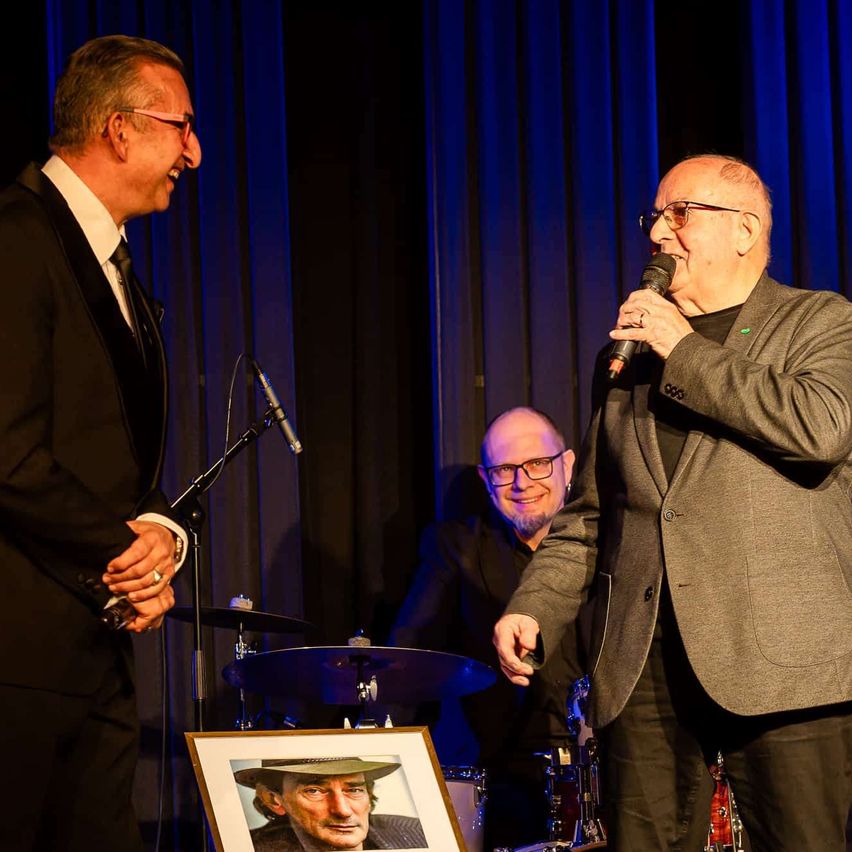 Zwei Männer in Anzügen auf der Bühne; einer spricht in ein Mikrofon, während der andere lächelt zusieht. Hinter ihnen steht ein Porträt eines Mannes in einem Hut. Musikinstrumente, einschließlich Trommeln und Becken, sind vorhanden.