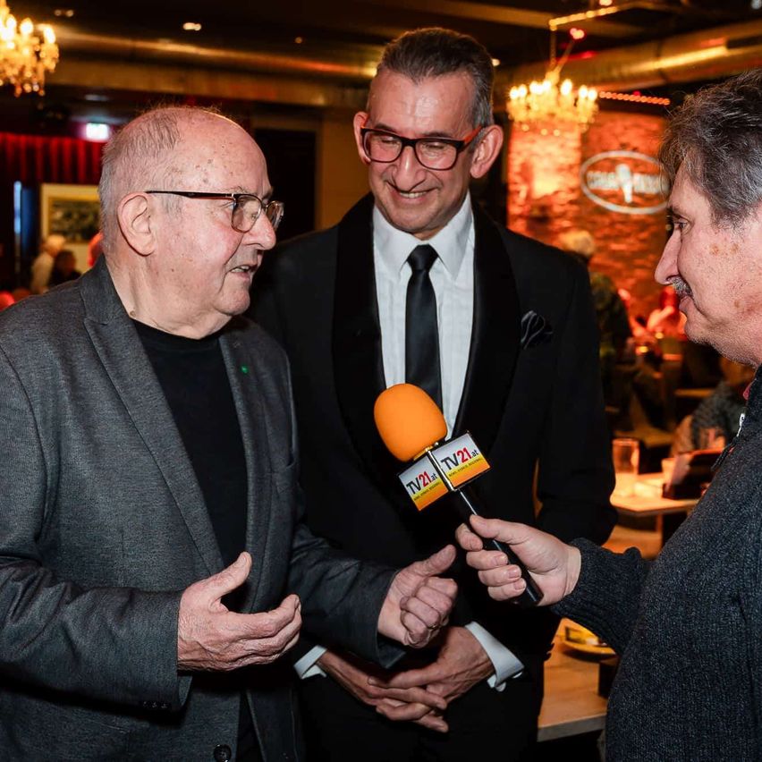 Ein älterer Mann in einem Anzug wird von einer Person mit einem Mikrofon mit dem TV21-Logo interviewt. Hinter ihm steht ein Mann in einem schwarzen Anzug mit Krawatte und lächelt. Im Hintergrund befindet sich ein roter Vorhang und eine Kronleuchter.