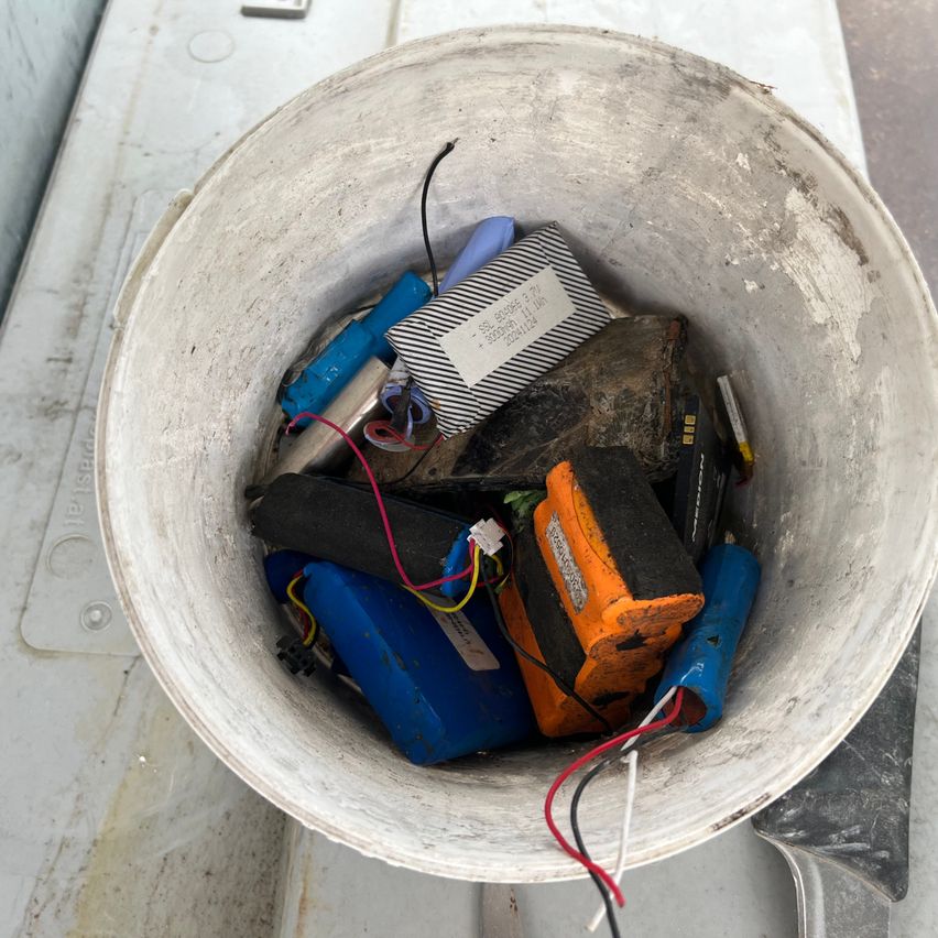 A white bucket filled with various electronic components including batteries, wires, and a small box. The bucket is placed on a concrete surface.