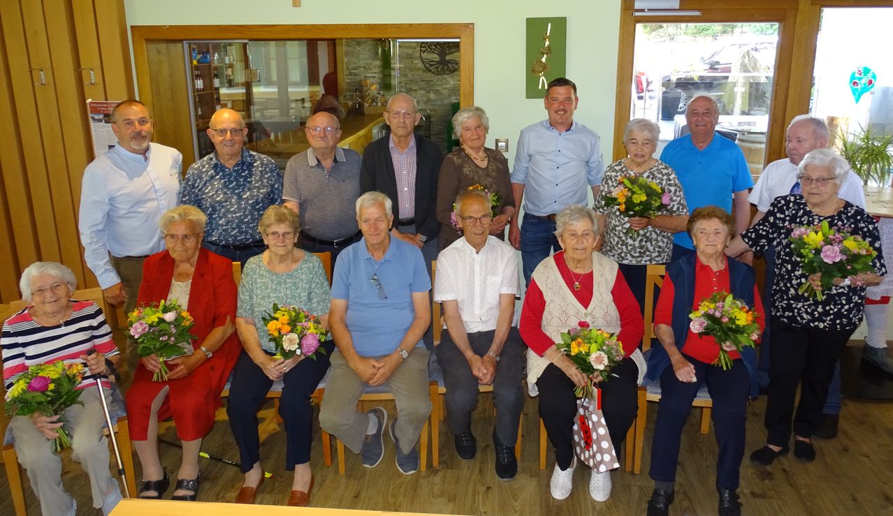 Eine Gruppe älterer Personen, in verschiedenen Kleidungsstücken gekleidet, sitzt gemeinsam auf Stühlen, lächelt und hält Blumen. Sie sind drinnen versammelt.