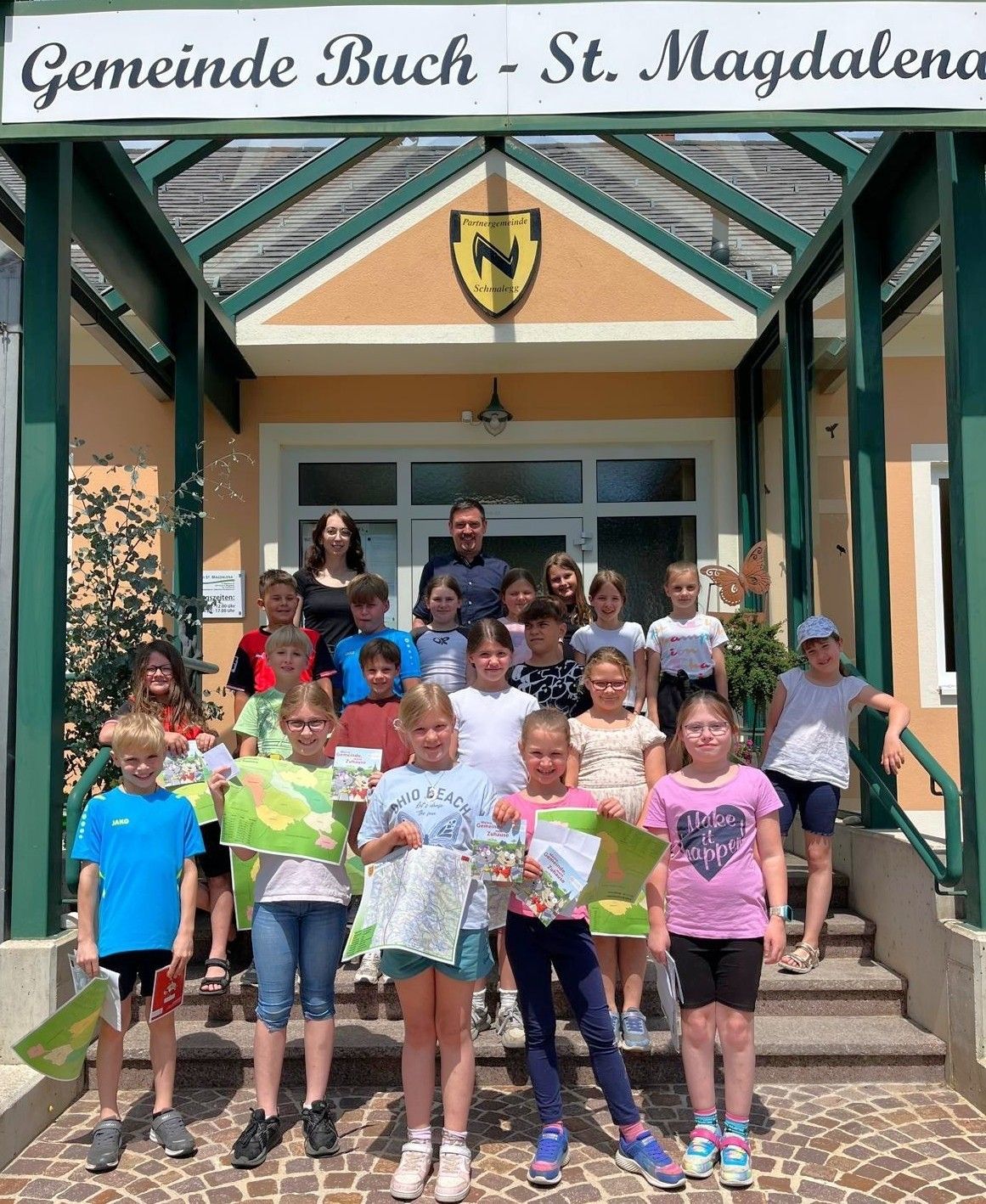 Eine Gruppe von Kindern und Erwachsenen steht vor einem Gebäude auf Stufen und hält Bücher und Karten. Ein Mann und eine Frau stehen in der Mitte.