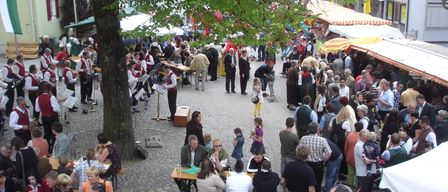 Ein überfüllter Außenmarkt mit Menschen, die um Holztische unter Sonnenschirmen sitzen. Eine Band spielt Musik im Hintergrund. Einige Personen tragen Kinder. Die Umgebung ist lebhaft mit verschiedenen Händlern und Teilnehmern.