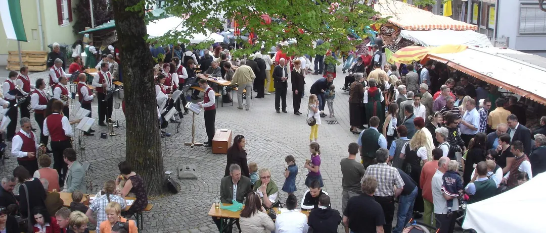 Ein überfüllter Außenmarkt mit Menschen, die um Holztische unter Sonnenschirmen sitzen. Eine Band spielt Musik im Hintergrund. Einige Personen tragen Kinder. Die Umgebung ist lebhaft mit verschiedenen Händlern und Teilnehmern.