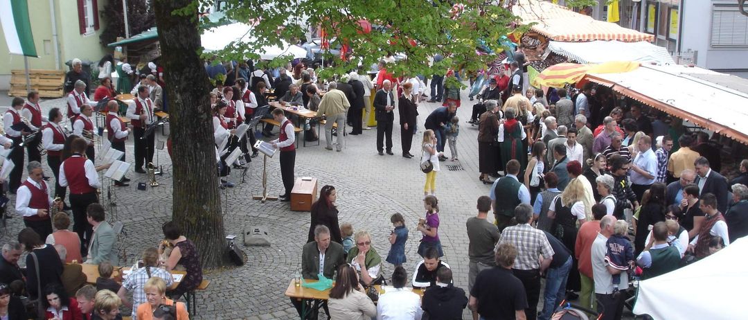 Ein überfüllter Außenmarkt mit Menschen, die um Holztische unter Sonnenschirmen sitzen. Eine Band spielt Musik im Hintergrund. Einige Personen tragen Kinder. Die Umgebung ist lebhaft mit verschiedenen Händlern und Teilnehmern.