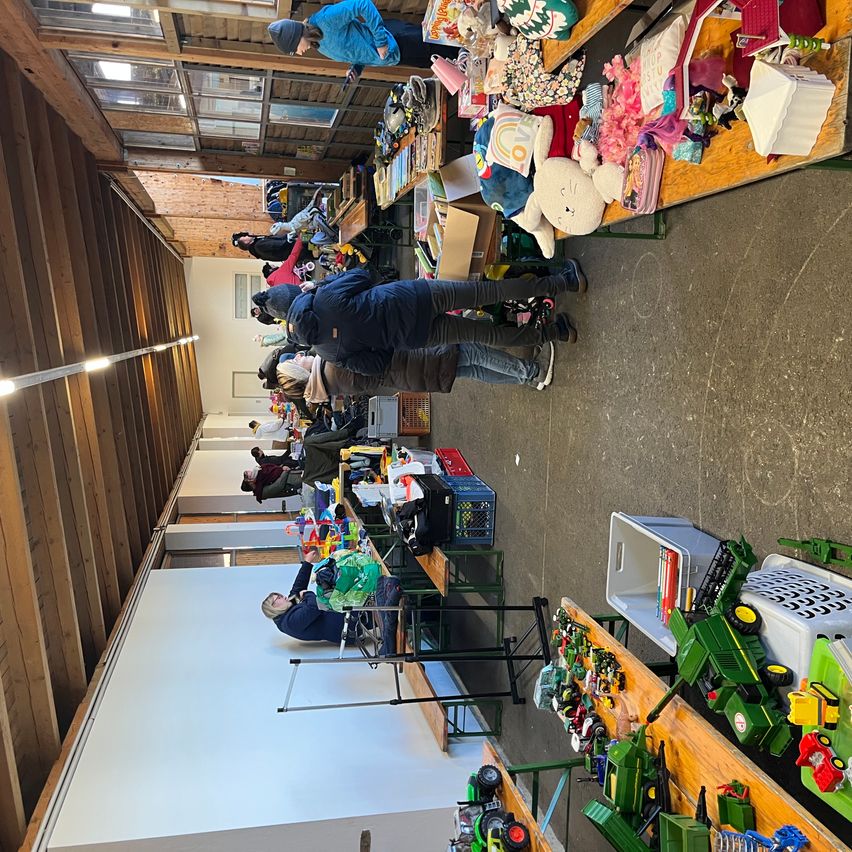 Ein Innenbereich mit vielen Spielzeugen und Gegenständen, die auf Tischen ausgestellt sind. Leute stöbern durch die Artikel. Holzstrukturen befinden sich an den Seiten.