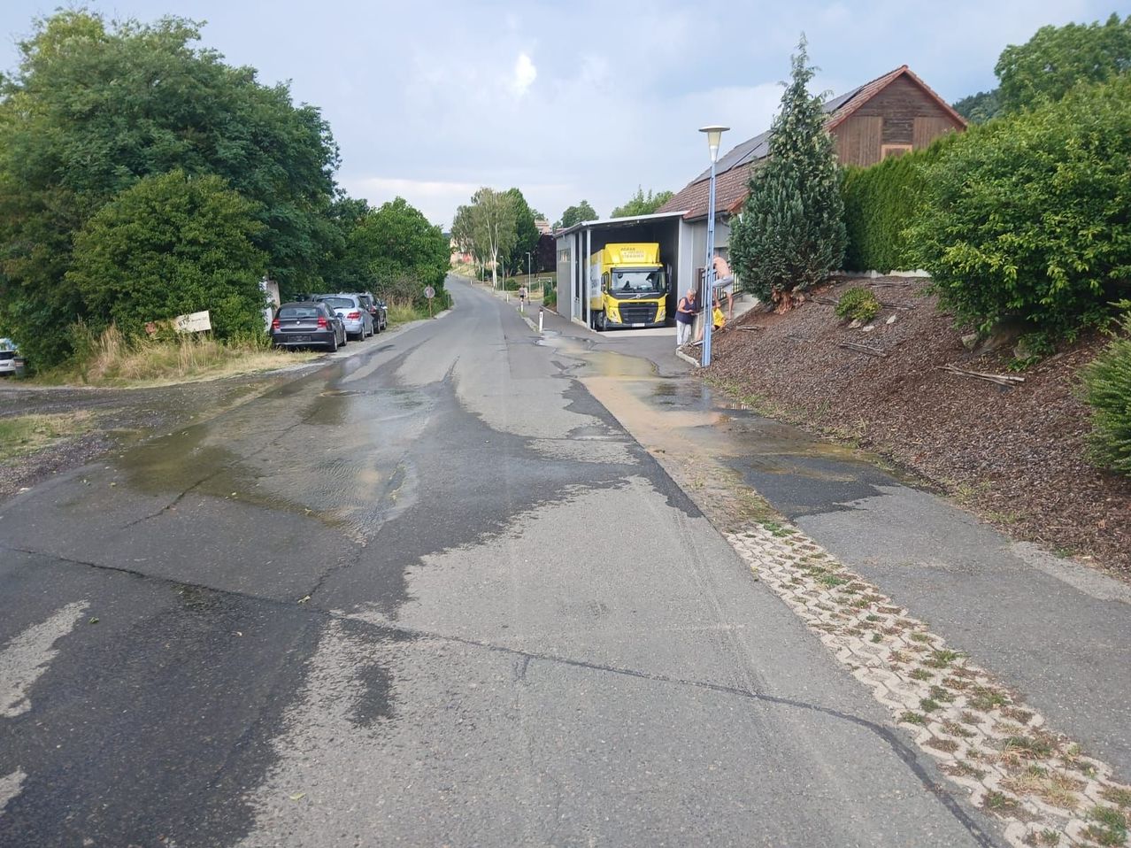Eine nasse Straße mit geparkten Autos, einem gelben Lkw und einer Person, die neben dem Gebäude mit Solarpaneelen auf dem Dach steht.