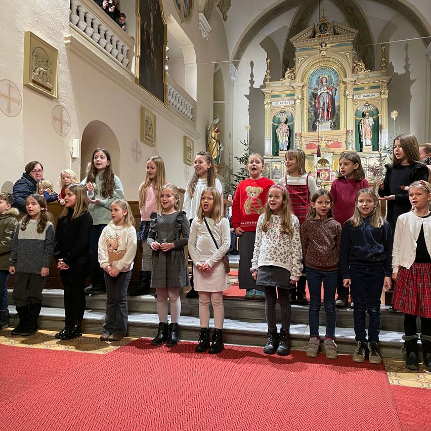 Eine Gruppe von Kindern und Erwachsenen steht auf einem roten Teppich in einer Kirche und singt. Sie tragen Winterkleidung. Hinter ihnen steht ein Weihnachtsbaum und ein Altar.