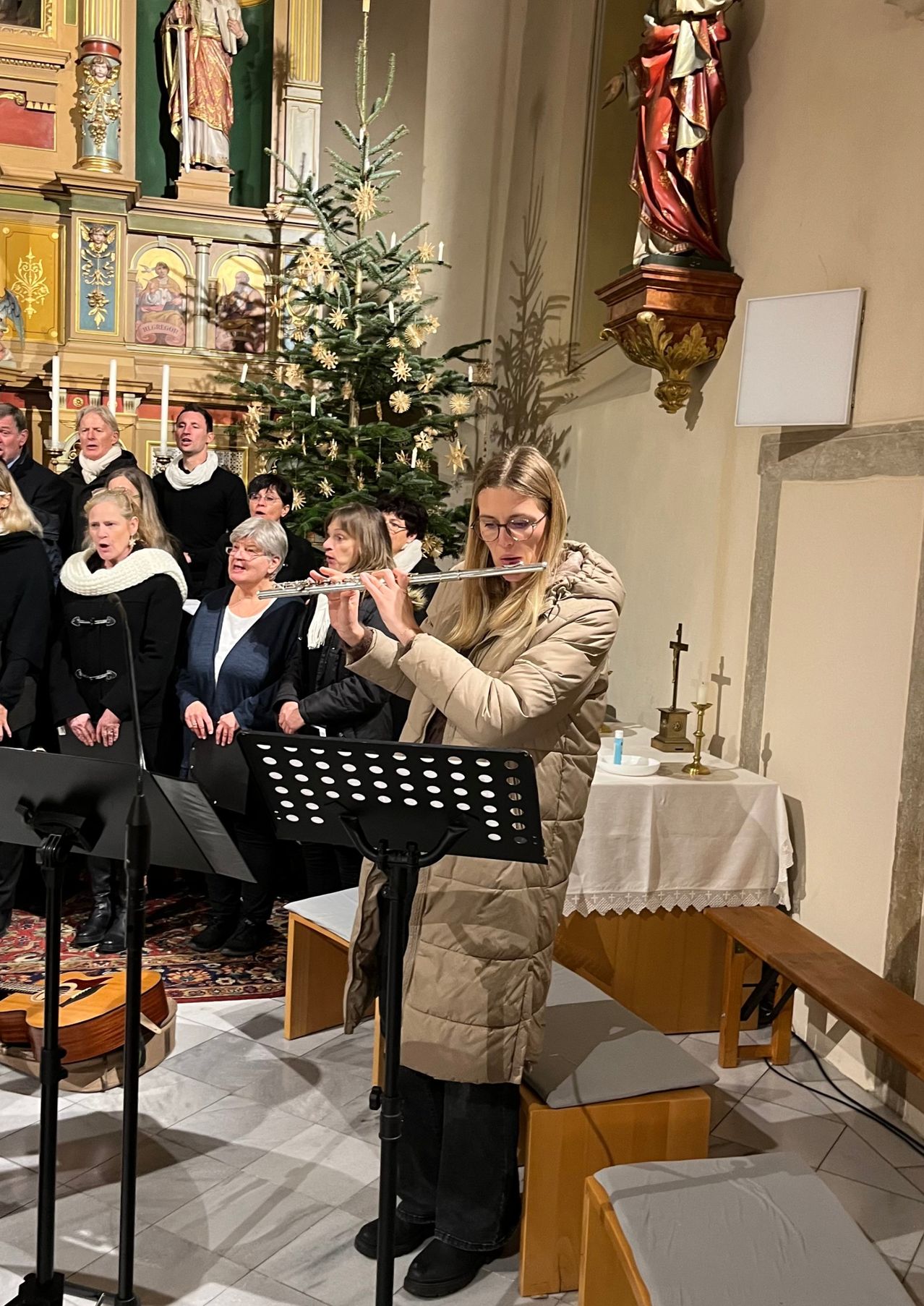 Eine Frau spielt Flöte, während ein Chor in einer Kirche singt, mit einem Weihnachtsbaum und einer Kerze im Hintergrund.