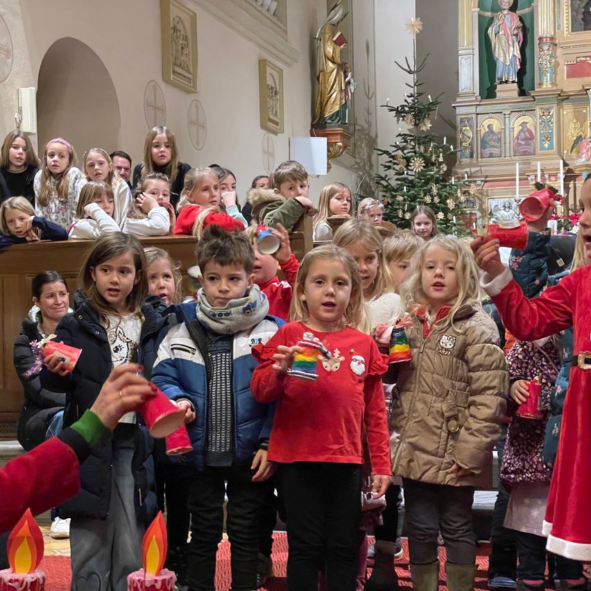 Kinder in Winterkleidung und roten Kleidern stehen in einer Kirche, halten Tassen und Kerzen. Sie schauen zu einer Frau, die eine Tasse hält, mit Erwachsenen und einem Weihnachtsbaum im Hintergrund.