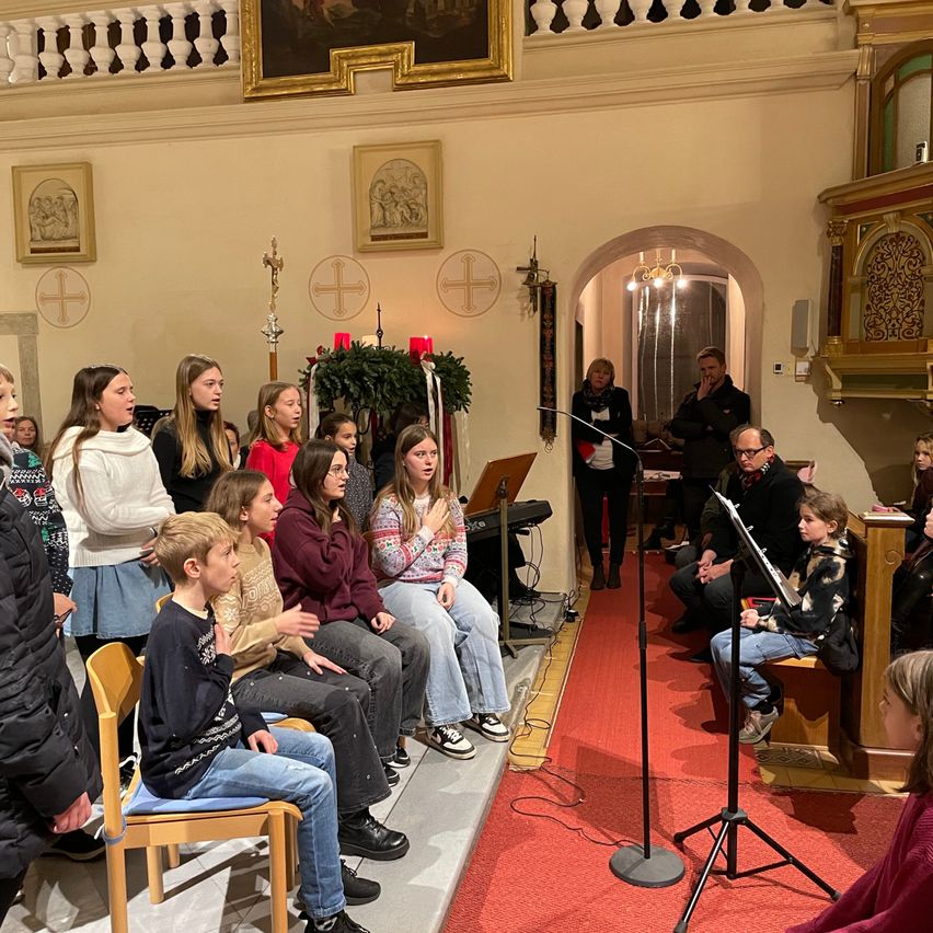 Ein Kinderchor singt in einer Kirche mit Klavier und Mikrofonen auf der Bühne. Die Leute sitzen auf Kirchenbänken und Stühlen auf beiden Seiten.