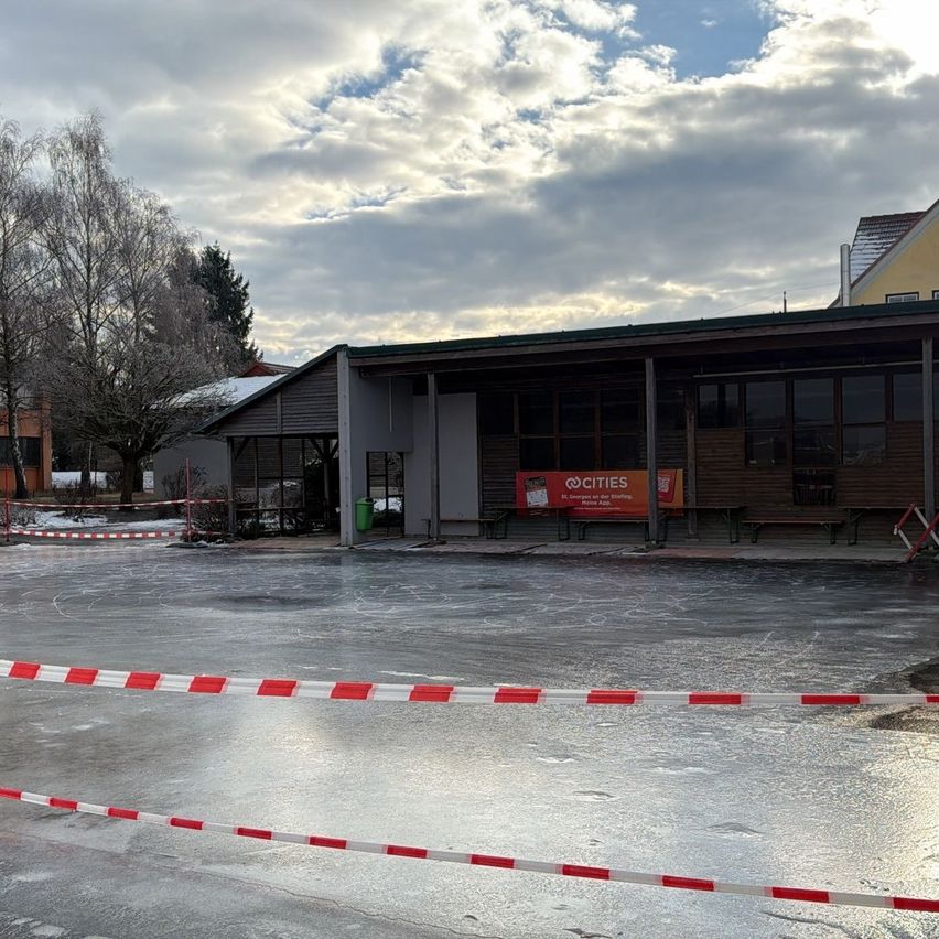 Ein leerer, vereister Bereich mit einer roten und weißen Barriere davor, mit einem Gebäude, an dessen Wand ein Schild mit der Aufschrift '@CITIES' steht.