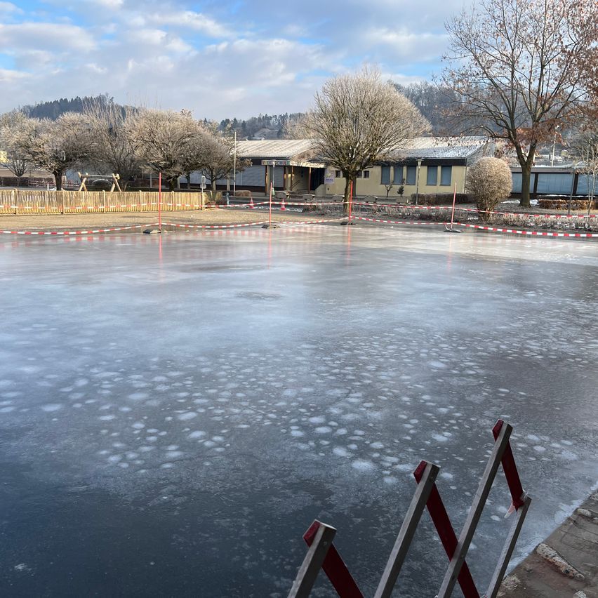 Eine Außeneisbahn mit einem gelben Gebäude im Hintergrund. Die Bahn ist von einem Zaun mit roten und weißen Streifen umgeben. Bäume und Pflanzen befinden sich rund um die Bahn.