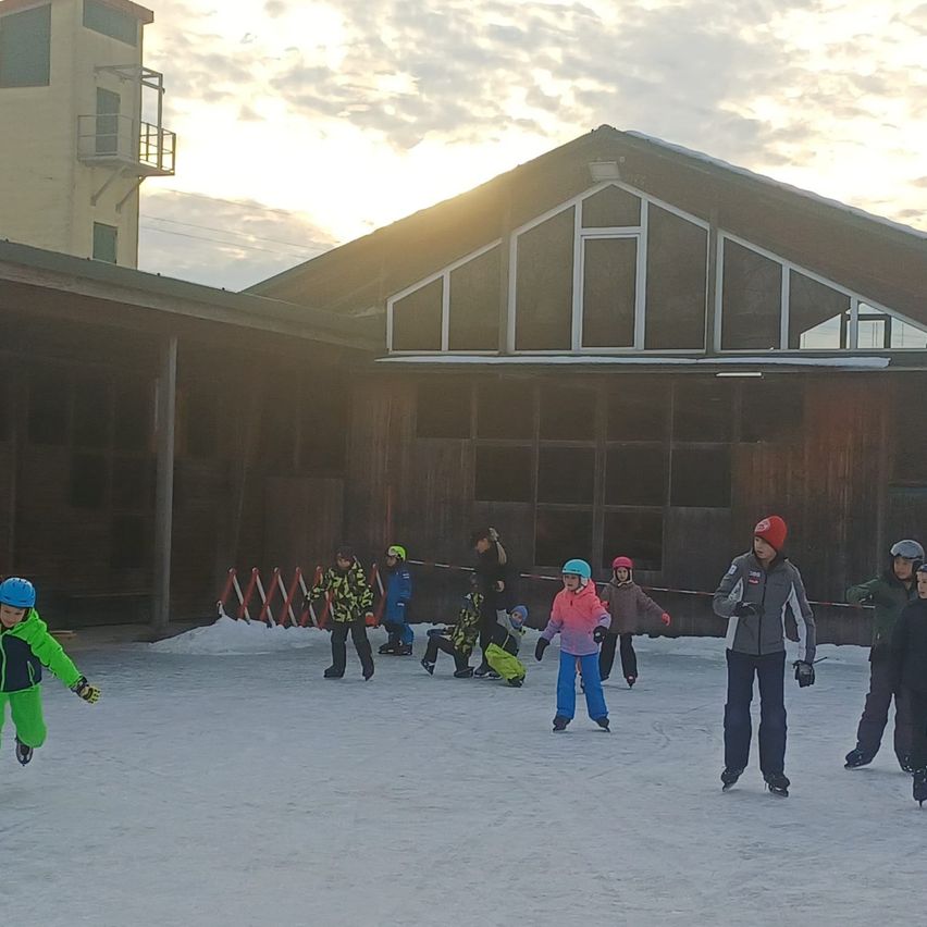 Eine Gruppe von Kindern spielt Eishockey auf einer Eisbahn und trägt Helme und Handschuhe. Hinter ihnen befindet sich ein Gebäude mit Glasfenstern und einem Zaun.