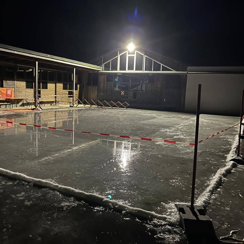 Eine beleuchtete Eisbahn bei Nacht, beleuchtet von einem hellen Licht. Die Bahn ist mit rot-weißem Band eingezäunt.