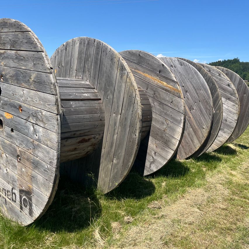 Eine Reihe verwitterter Holzspulen für Kabel liegt auf einem Grasfeld mit blauem Himmel im Hintergrund.