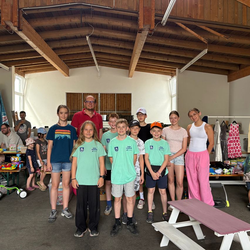 Eine Gruppe junger Leute in gleichen grünen T-Shirts steht in einem Raum mit Holzbalken, einige mit Mützen. Dahinter posiert ein Mann in einem roten Hemd mit einem Mädchen. Andere Menschen, einige mit Mützen, sind in der Nähe von Tischen mit Kleidung und Spielzeug.