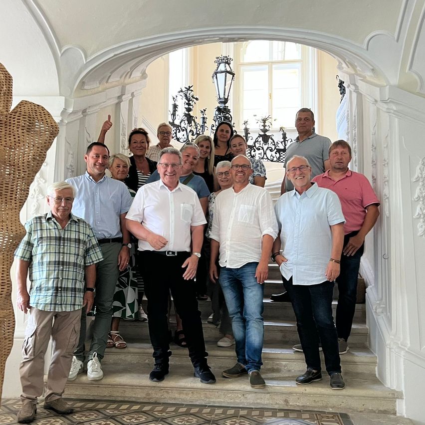 Eine Gruppe Erwachsener, einige in formeller Kleidung, stehen auf einer Treppe in einem prächtigen Raum mit gewölbten Türen und Kronleuchtern. Sie posieren für ein Foto.