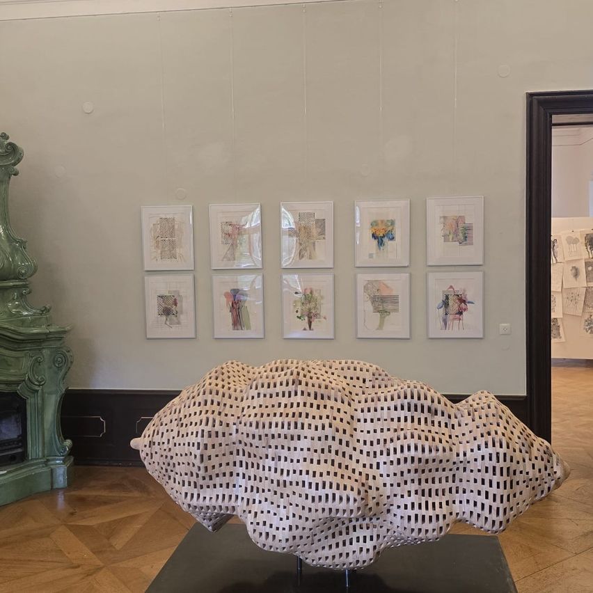 Eine Kunstgalerie zeigt eine große, strukturierte Skulptur auf einem schwarzen Podest und einen Kamin. Darüber befinden sich gerahmte Zeichnungen an der Wand.
