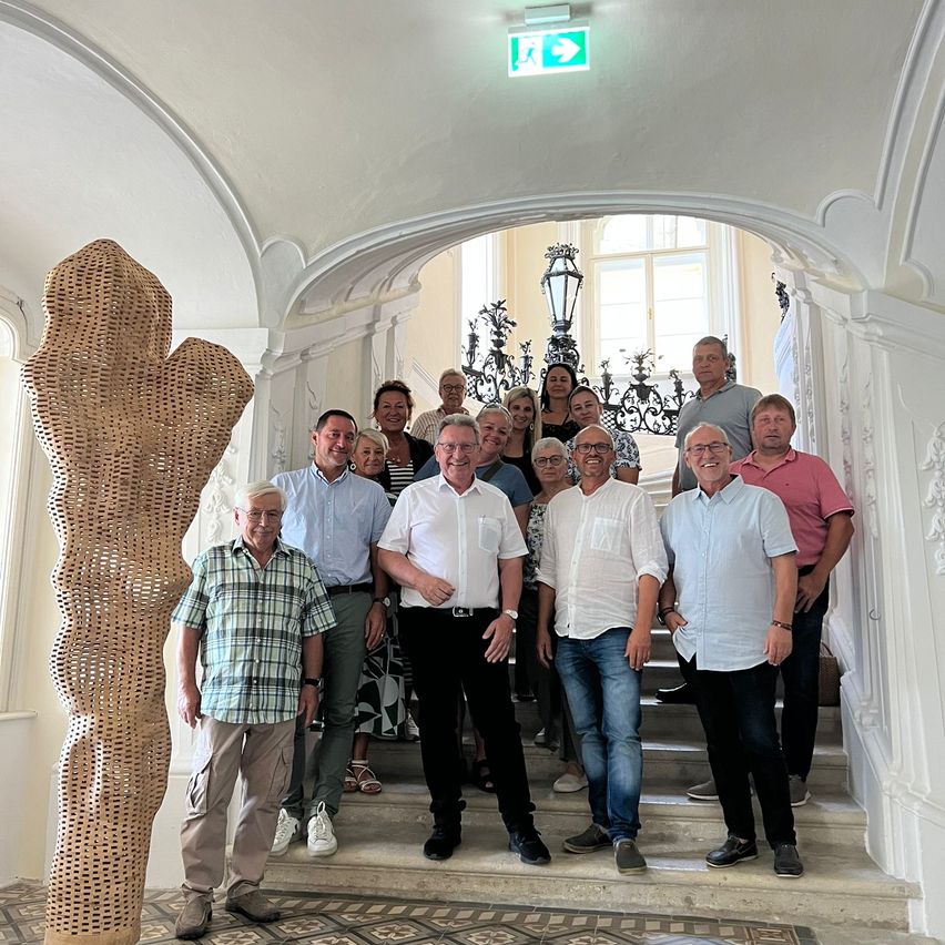 Eine Gruppe von Erwachsenen steht in einem kunstvoll gestalteten Flur mit einer großen Korbskulptur und posiert für ein Foto. Sie tragen Freizeitkleidung, und der Flur ist mit weißen Wänden und dekorativen Elementen geschmückt.