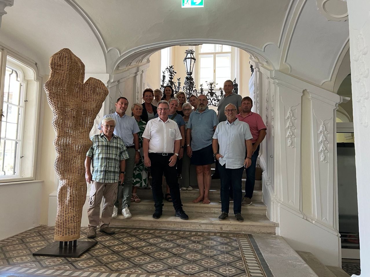 Eine Gruppe von Menschen steht in einem Flur mit einer großen Weidenskulptur und posiert für ein Foto. Sie sind lässig gekleidet. Der Flur weist weiße Wände und dekorative Muster auf.