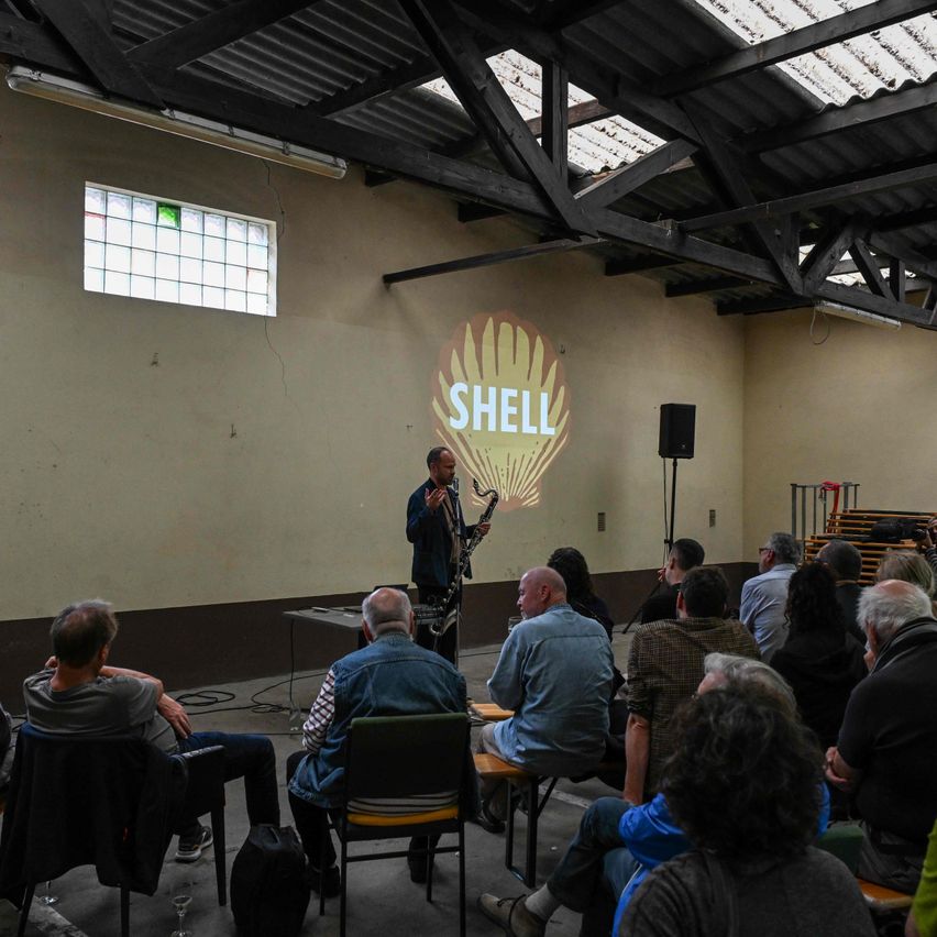 Ein Mann spielt Saxophon vor einem Publikum in einem Raum mit Shell-Logo an der Wand.