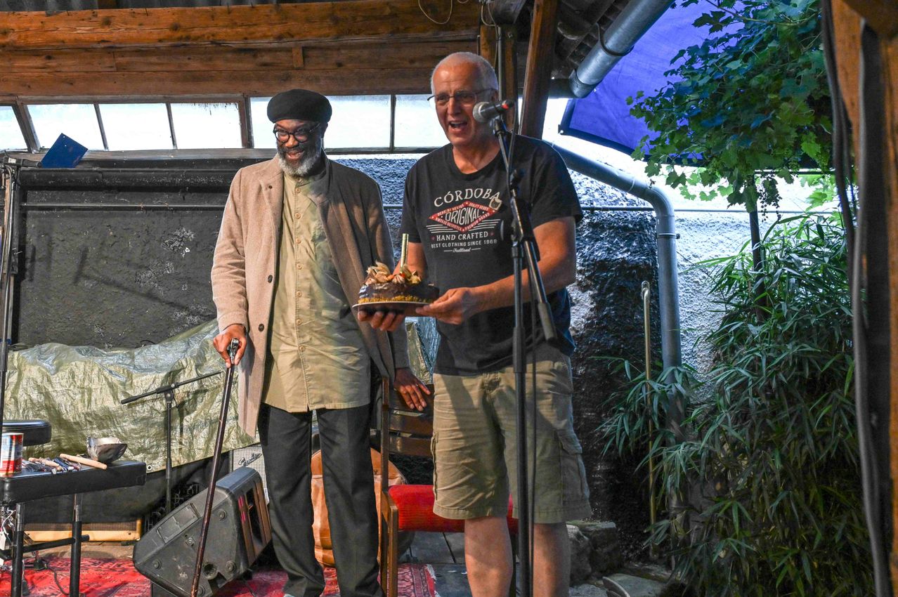 Zwei Männer stehen vor einem Mikrofon. Einer hält einen Kuchen, während der andere lächelt und spricht. Dahinter sind ein Lautsprecher und eine Pflanze zu sehen.