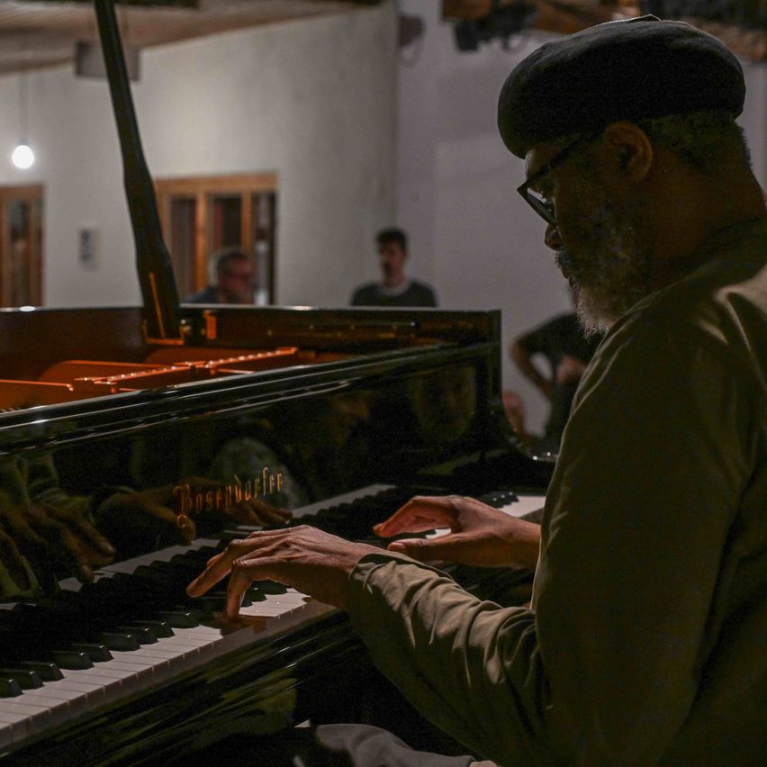Ein Mann spielt mit ernster Miene das Klavier in einem schwach beleuchteten Raum. Ein anderer Mann beobachtet ihn von hinten.