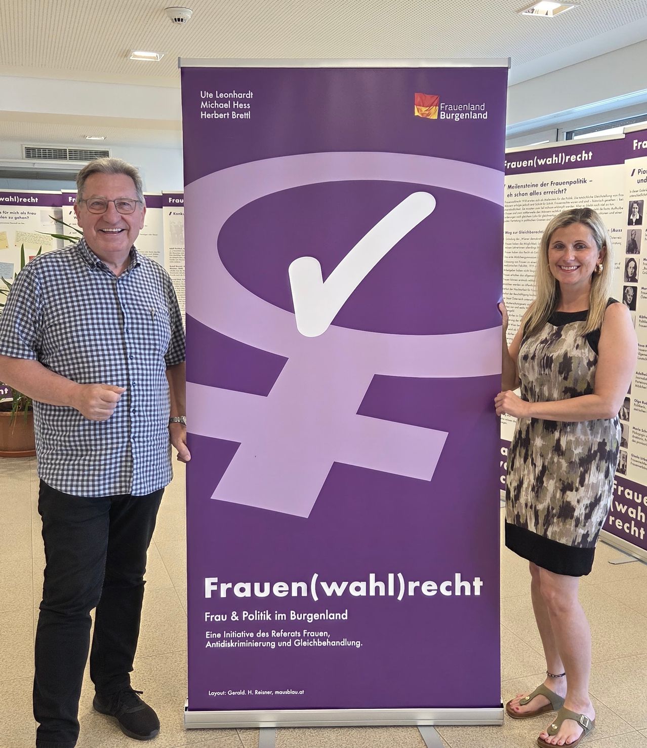 Zwei Personen stehen neben einem violetten Plakat mit einem weißen Haken und Text. Ein Mann trägt ein kariertes Hemd und schwarze Hose, und eine Frau trägt ein schwarzes und weißes Kleid. Sie lächeln beide.