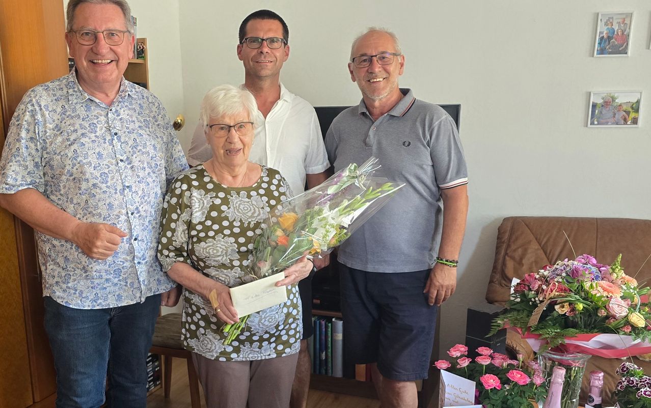 Eine ältere Frau steht mit einem Blumenstrauß in der Hand, umgeben von drei lächelnden Männern in einem Raum. Dahinter befindet sich ein Regal mit Büchern und einem Fernseher.