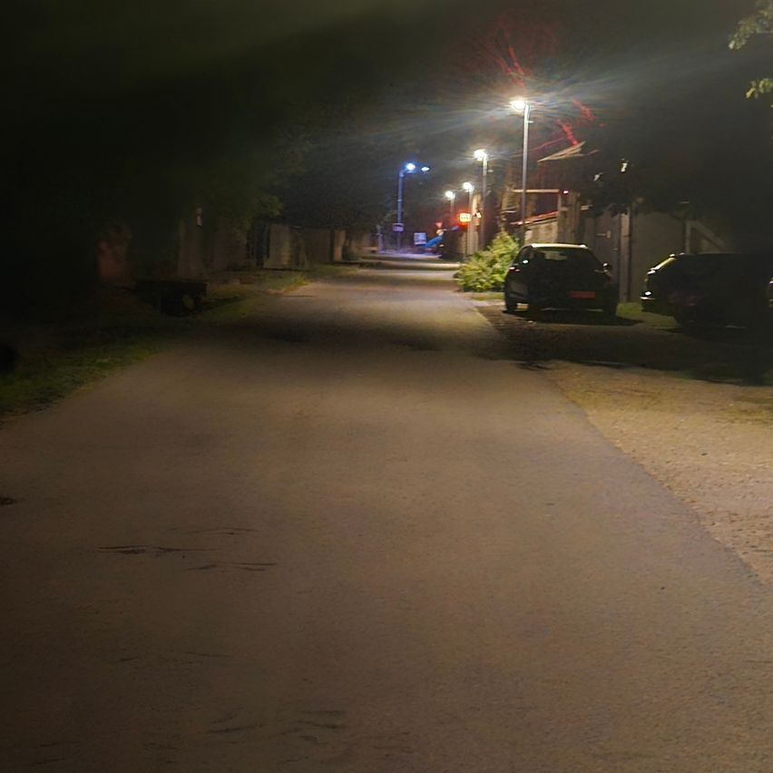 Eine Nachtaufnahme einer Straße mit zwei geparkten Autos auf der rechten Seite, beleuchtet durch Straßenlaternen.