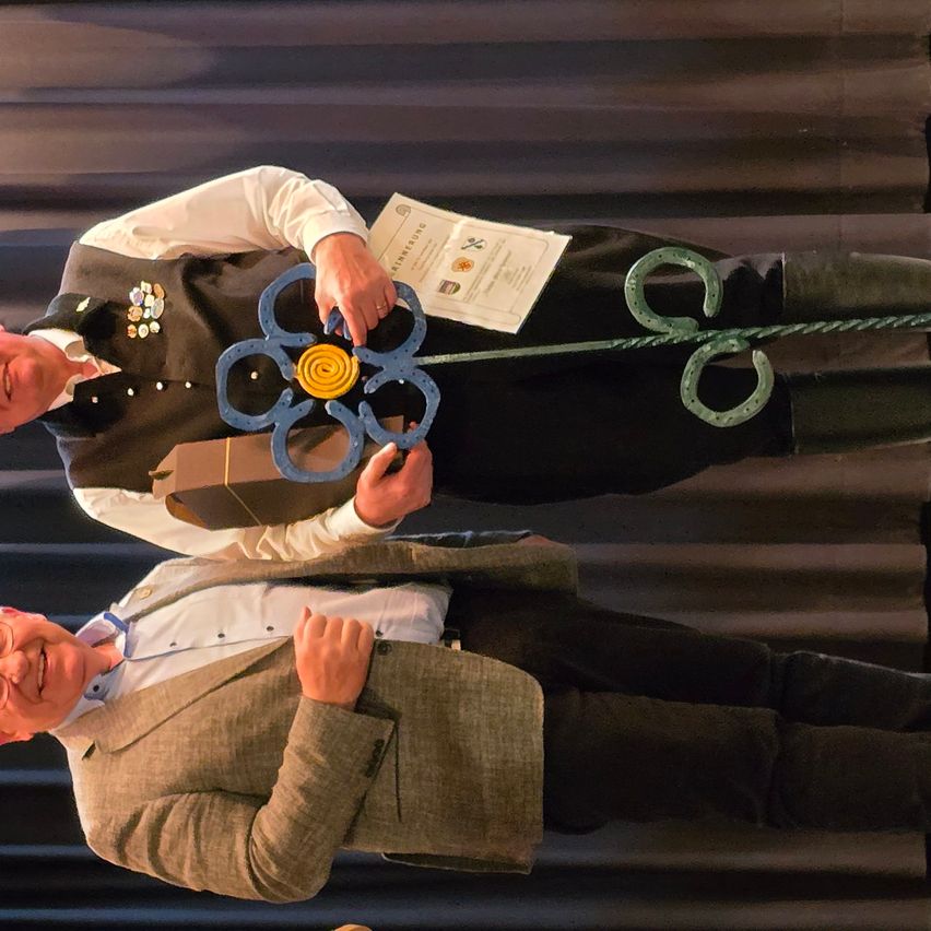 Zwei Männer stehen mit einem blumenförmigen Objekt zwischen ihnen, einer hält es und der andere zeigt den Daumen nach oben. Sie sind formell gekleidet, und einer hält ein Zertifikat.