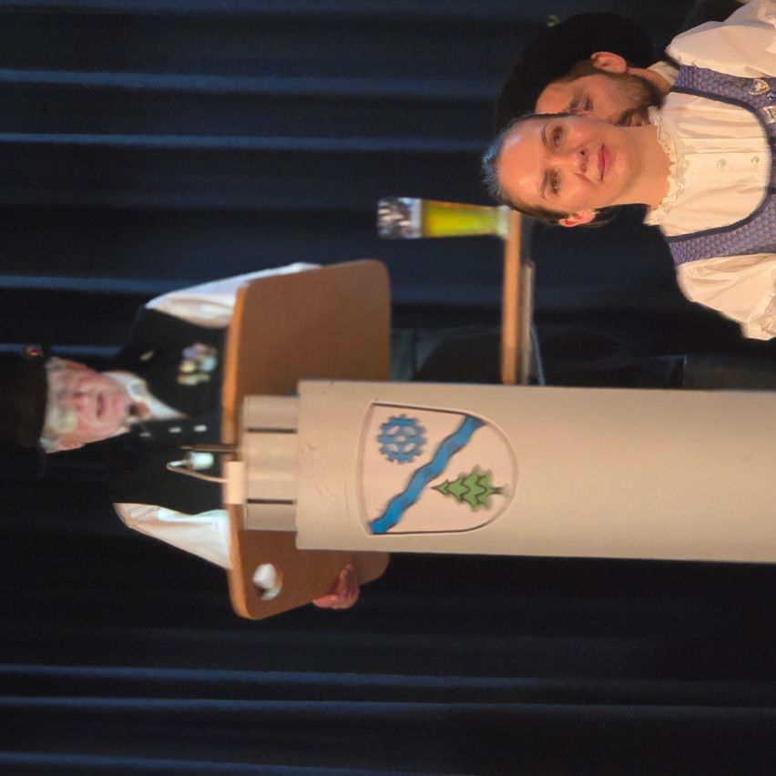 Ein Mann in traditioneller Kleidung spricht an einem Podium, während eine Frau zusieht und ein großes Emblem hinter ihnen steht.