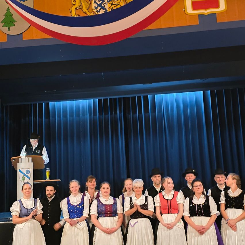 Eine Gruppe von Erwachsenen in traditionellen Kostümen steht auf der Bühne. Ein Mann steht hinter einem Podium, während ein großer Banner über der Bühne hängt.