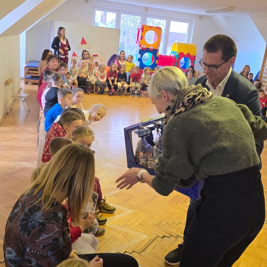 Eine Gruppe von Kindern und Erwachsenen versammelt sich in einem Raum. Eine erwachsene Frau hält eine Box mit Gegenständen und verteilt sie.