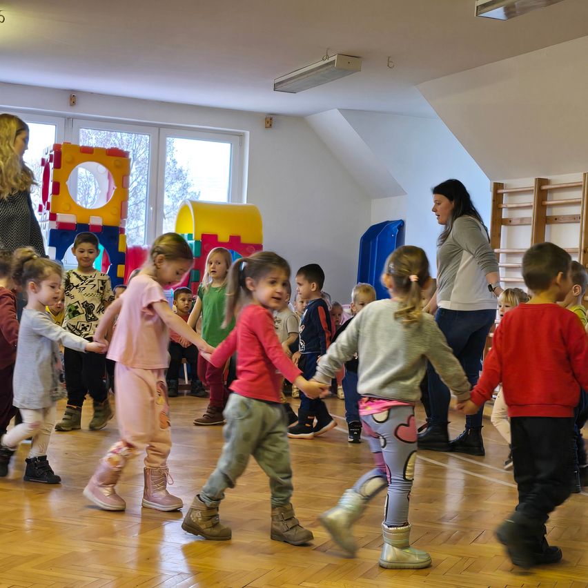 Eine Gruppe von Kindern hält sich an den Händen und tanzt in einem Raum mit Spielzeug und einer Lehrerin.