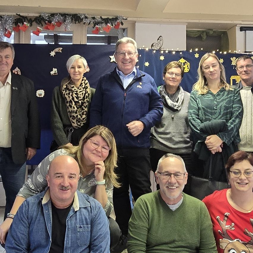 Eine Gruppe von Menschen posiert für ein Foto in einem Raum, der mit blauen Vorhängen und Weihnachtsdekorationen geschmückt ist. Die Menschen umfassen sowohl Männer als auch Frauen, einige tragen Brillen und Schals. Sie scheinen zu lächeln und für die Kamera zu posieren.