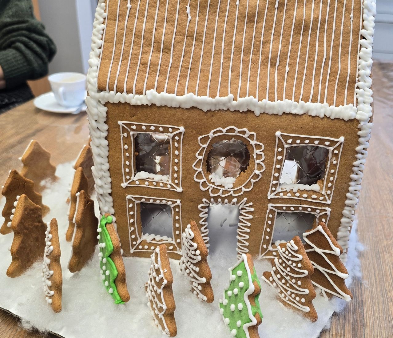 Ein Lebkuchenhaus mit kunstvoll gestalteten Fenstern und Tür, verziert mit weißer Glasur und grünen Ornamenten, steht auf einem Holztisch, mit einer Tasse Kaffee in der Nähe.
