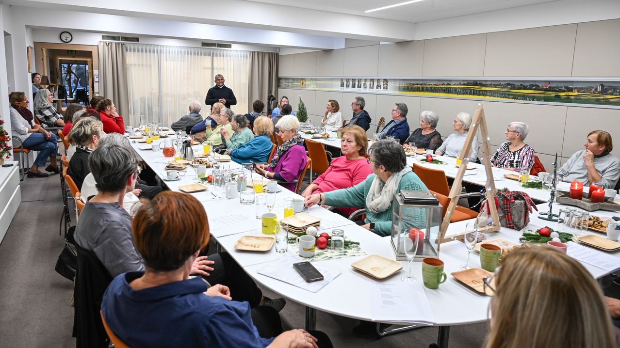 Eine Gruppe älterer Menschen sitzt um einen langen Tisch in einem Konferenzraum herum und genießt eine Mahlzeit. Sie werden von einem Mann begleitet, der in der Nähe des Fensters steht. Der Tisch ist mit verschiedenen Gerichten, Gläsern und Tellern geschmückt.