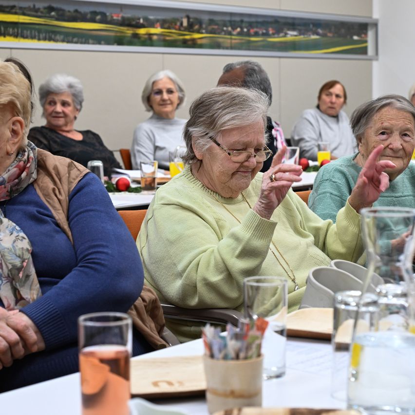Mehrere ältere Frauen sitzen um einen Tisch herum, möglicherweise in einer Besprechung, mit Gläsern und einem Teller auf dem Tisch. Die Frau in der Mitte trägt eine Brille und gestikuliert. Hinter ihnen eine Wand mit einem großen Gemälde.