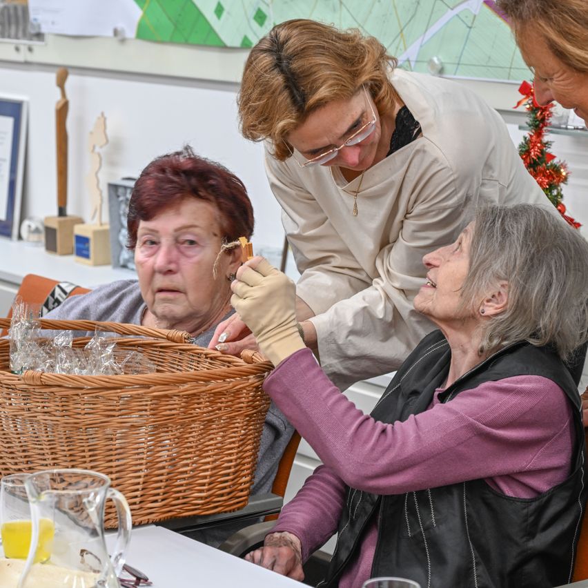 Drei Frauen sitzen, eine mit einem Korb voller Glasbecher. Eine Frau in einer weißen Jacke hält ein Sandwich und füttert die Frau im schwarzen Westchen. Eine weitere Frau schaut von hinten zu.