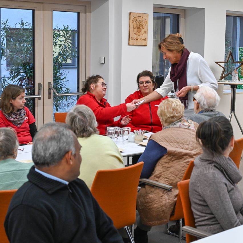 Eine Gruppe von Menschen sitzt in einem Raum mit einem Tisch davor. Eine Frau reicht einer anderen Frau ein Papier. Der Raum hat Glastüren und Fenster.