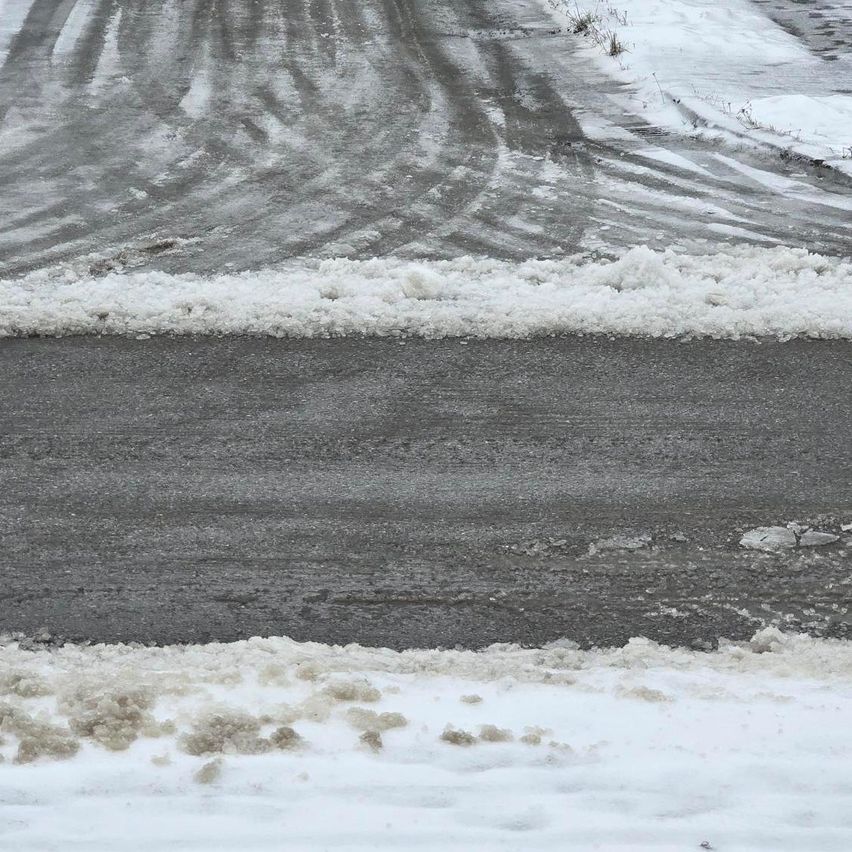 Schnee und Eis bedecken eine Straße, mit sichtbaren Fahrzeugspuren. Die Straße ist nass und erscheint eisglänzend. Schneebänke befinden sich auf beiden Seiten.