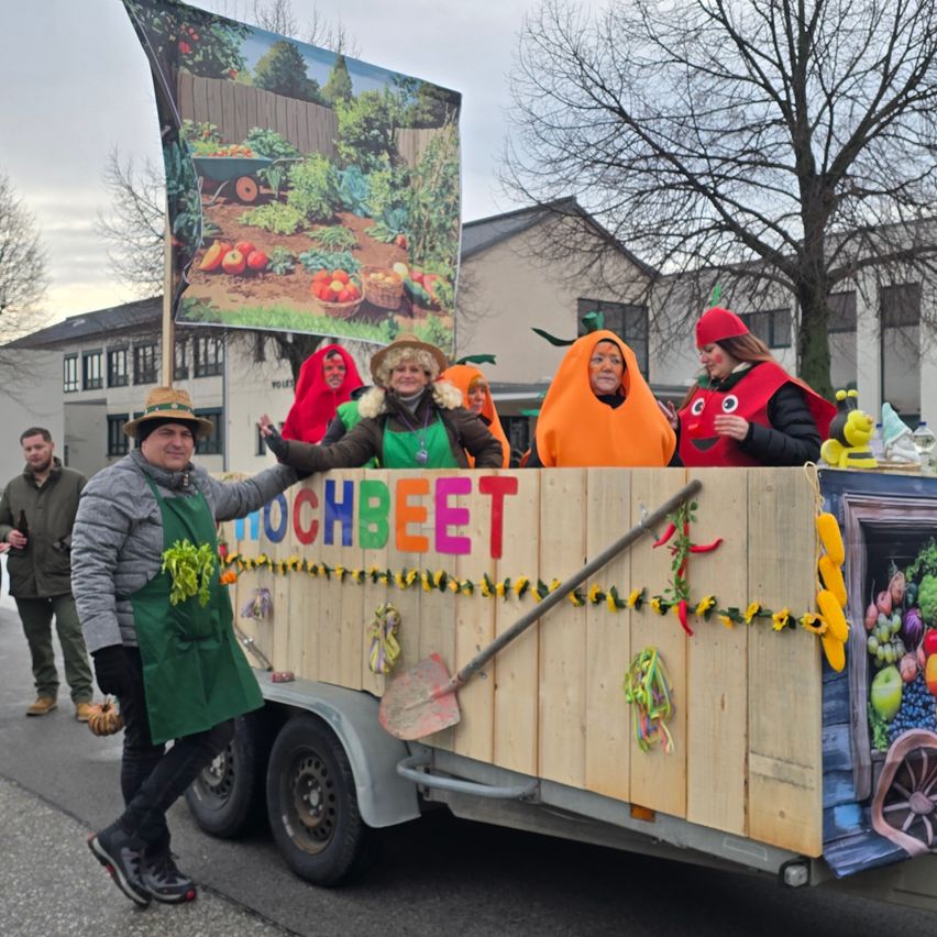 Menschen in Gemüsekostümen nehmen an einer Parade teil. Ein Wagen mit Gartenthema hat Menschen in Kürbis- und Rübenkostümen. Ein Mann hält eine Flagge mit einer Gartenszene.