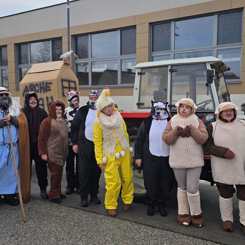 Eine Gruppe von Menschen in verschiedenen Kostümen, darunter Schafe, Hühner und ein Hirte, steht vor einem Gebäude mit großen Fenstern und einem Traktor.