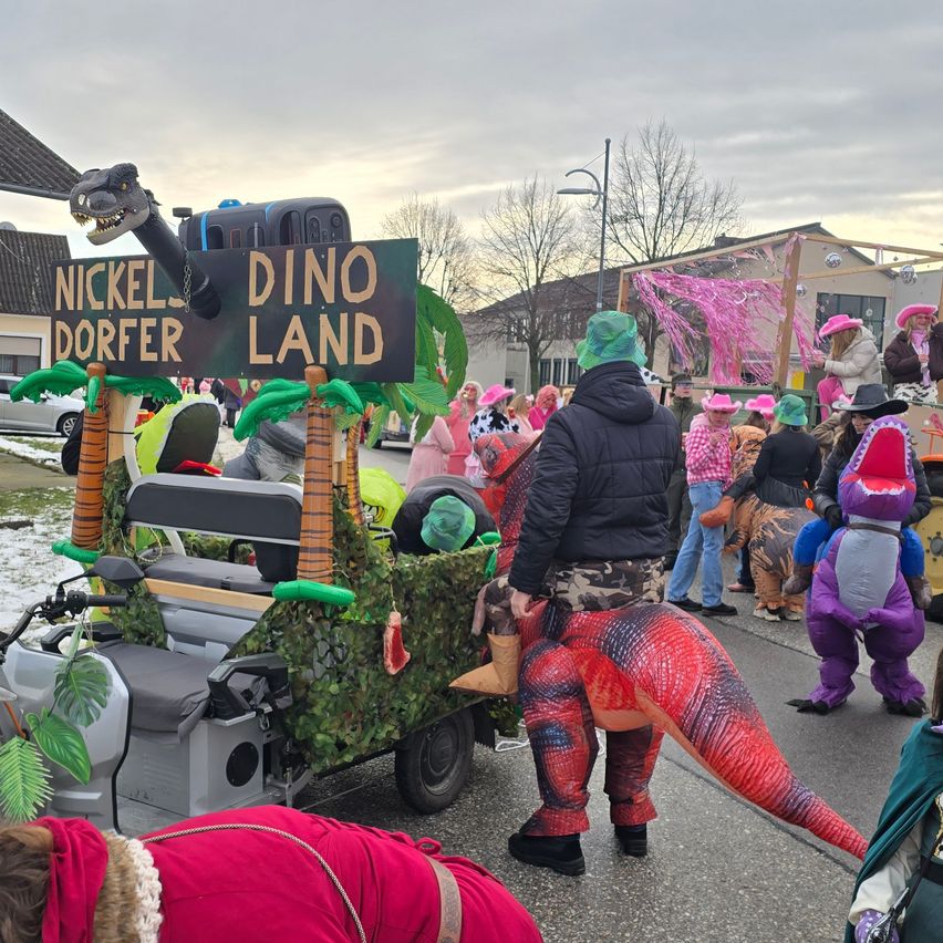 Ein Dinosaurier-Themen-Wagen ist bei einer Parade, mit Leuten in Dinosaurier-Kostümen und einer Person in einer schwarzen Jacke. Ein großes Schild sagt Nickel Dorfer Dino Land.