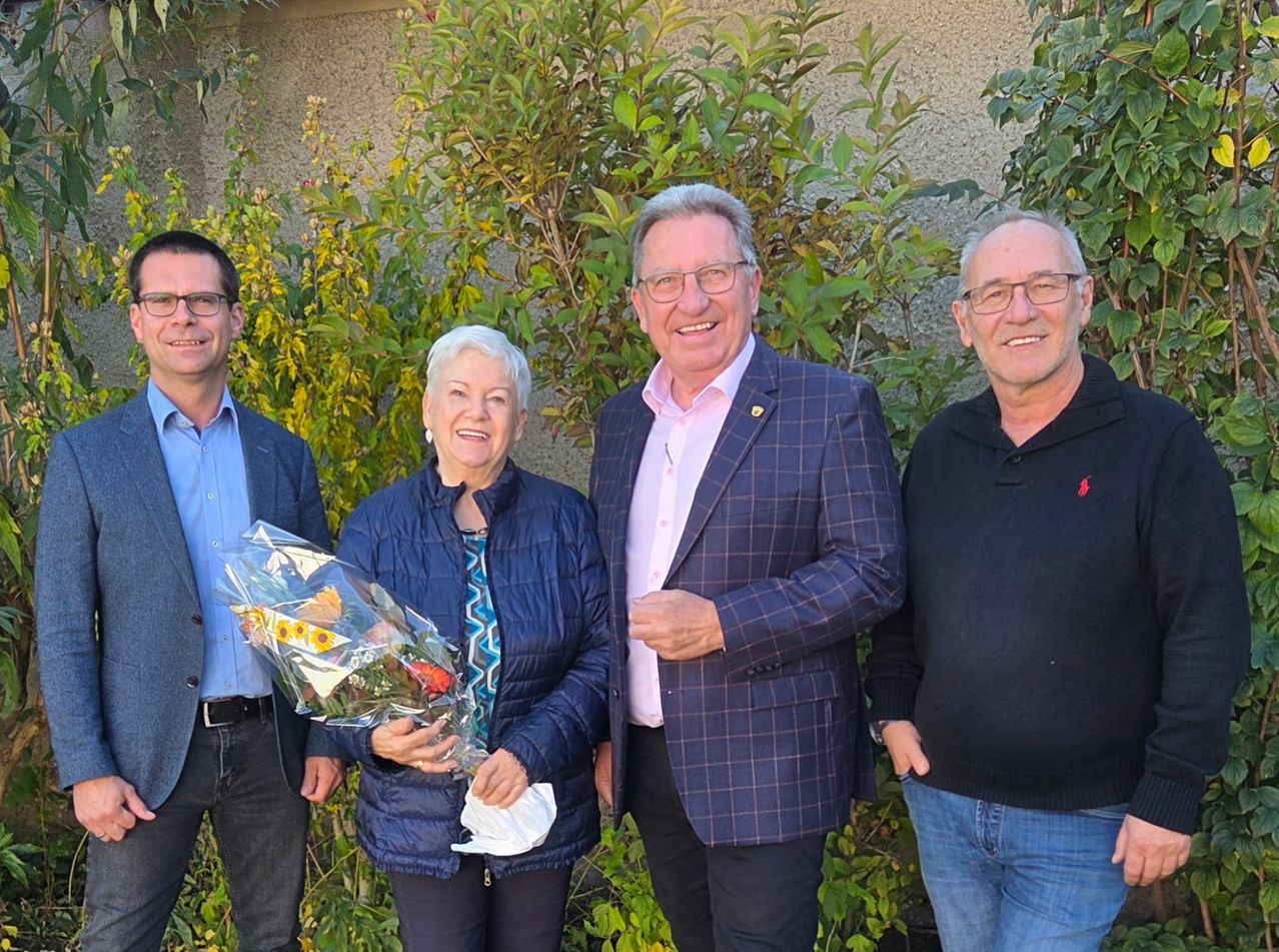 Four people stand close together in front of green bushes, smiling. A woman holds a bouquet of flowers.